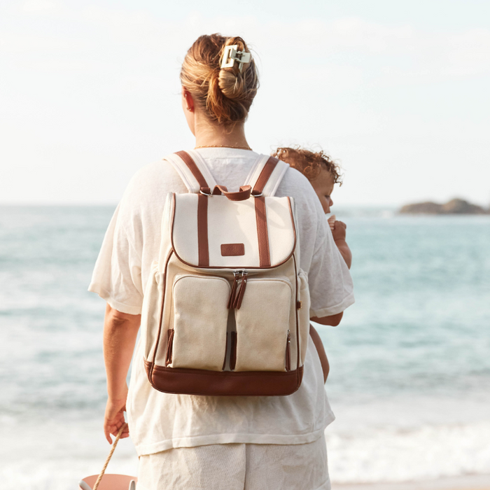 A mother wearing a backpack while carrying her baby along the beach, with the ocean in the background