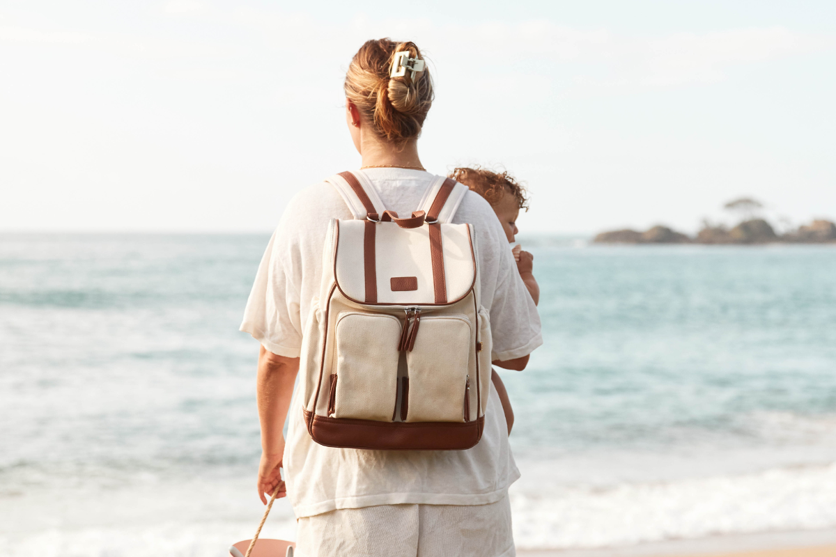A mother wearing a backpack while carrying her baby along the beach, with the ocean in the background