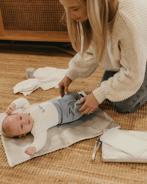 A mum getting ready to change her baby's nappy on the floor using a silver travel change mat.